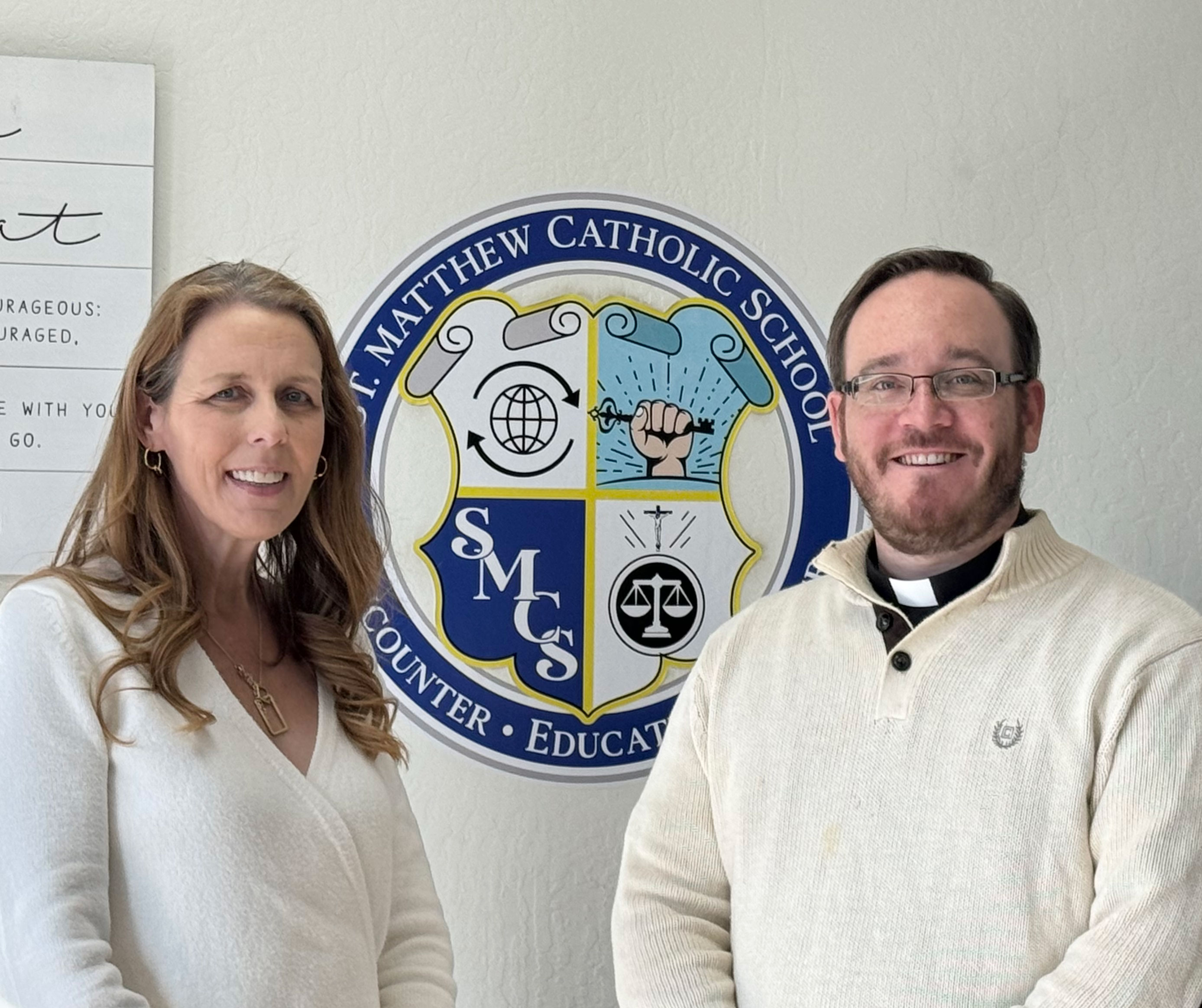 Principal Tax and Father Kevin standing in front of the school logo